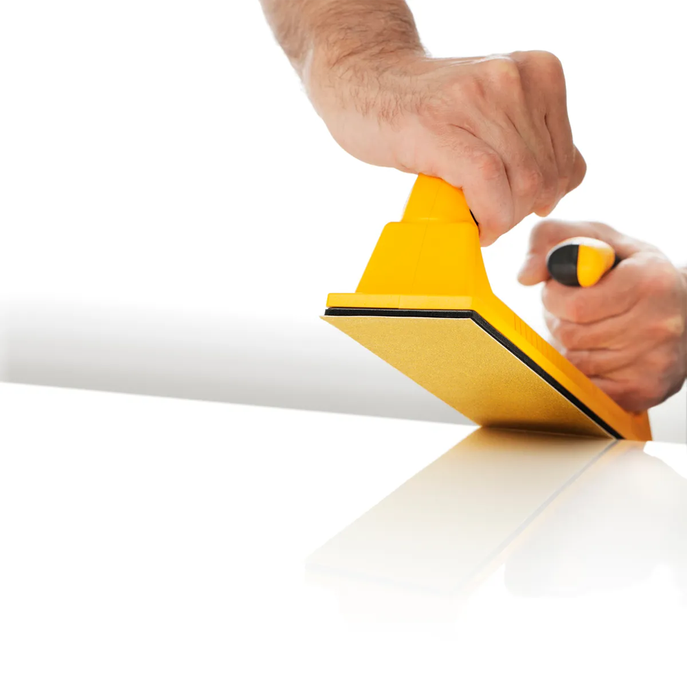 Hand using a yellow Mirka rigid board for precision manual sanding on a white panel