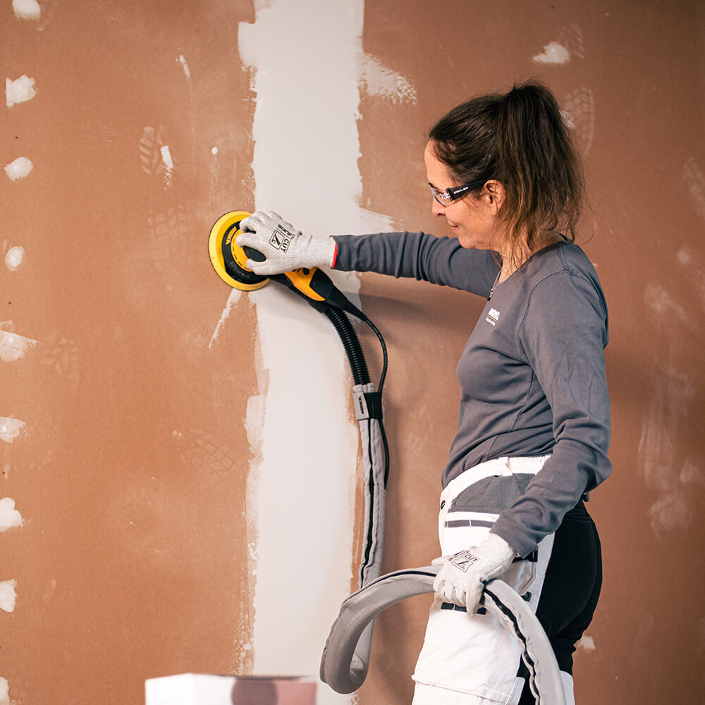 Female tradie using Mirka DEROS sander with dust extraction hose on a plasterboard wall